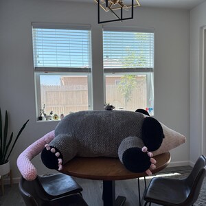 May include: A large, handmade crochet opossum plush toy in gray, black, and pink. The plush is lying on a wooden table, with its tail and paws visible. The opossum has black paws and ears, a pink tail, and a light-colored snout.