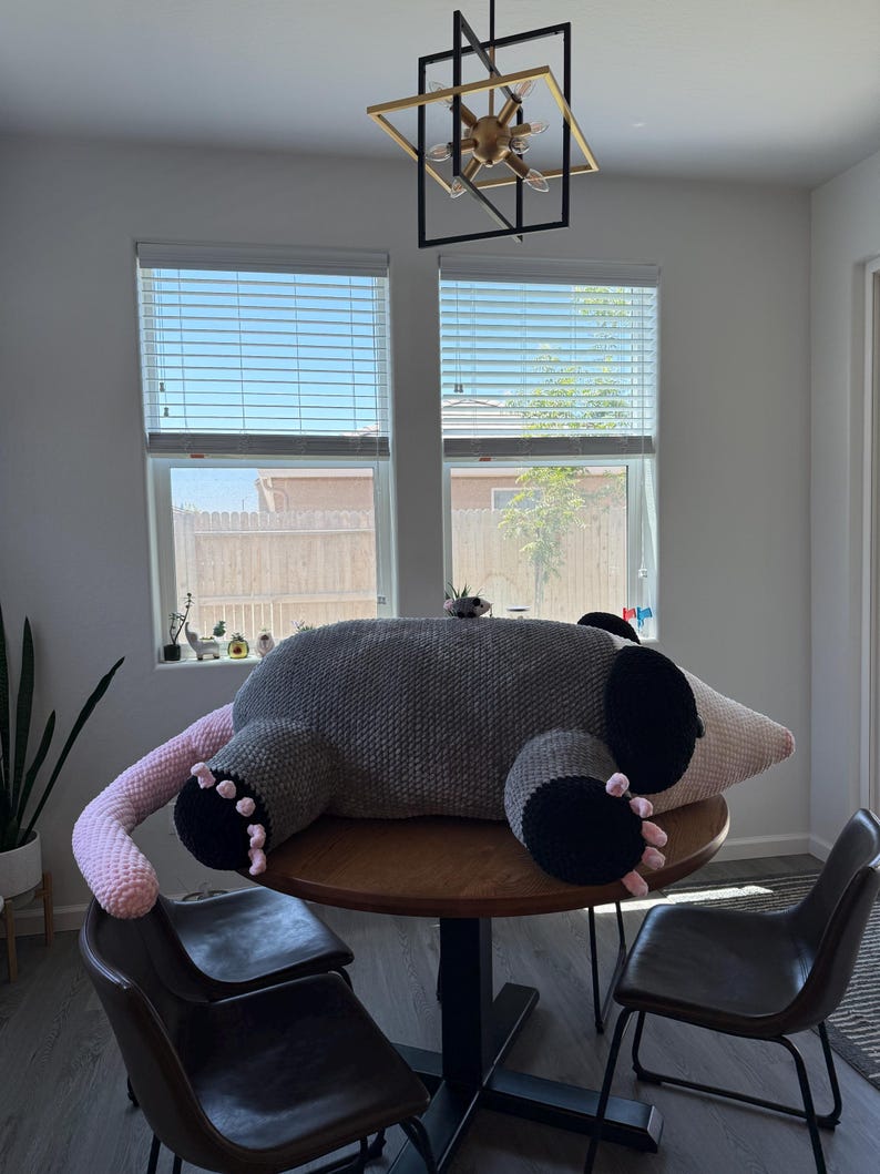 May include: A large, handmade crochet opossum plush toy in gray, black, and pink. The plush is lying on a wooden table, with its tail and paws visible. The opossum has black paws and ears, a pink tail, and a light-colored snout.