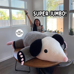 May include: A large, handmade crochet opossum plushie in shades of gray, white, black, and pink. The text "SUPER JUMBO!" is displayed above the plushie. The text "PDF PATTERN ONLY" is displayed on the left side of the image.