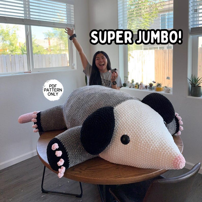 May include: A large, handmade crochet opossum plushie in shades of gray, white, black, and pink. The text "SUPER JUMBO!" is displayed above the plushie. The text "PDF PATTERN ONLY" is displayed on the left side of the image.