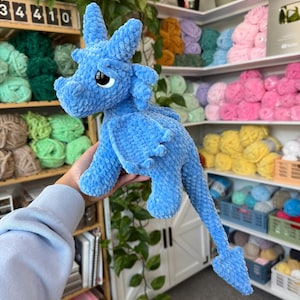 K&ouml;nnte beinhalten: Ein hellblauer geh&auml;kelter Drache mit gro&szlig;en schwarzen Augen, Fl&uuml;geln und einem langen Schwanz. Der Drache ist aus einem weichen, strukturierten Garn gefertigt. Der Hintergrund zeigt Regale voller bunter Wolle.