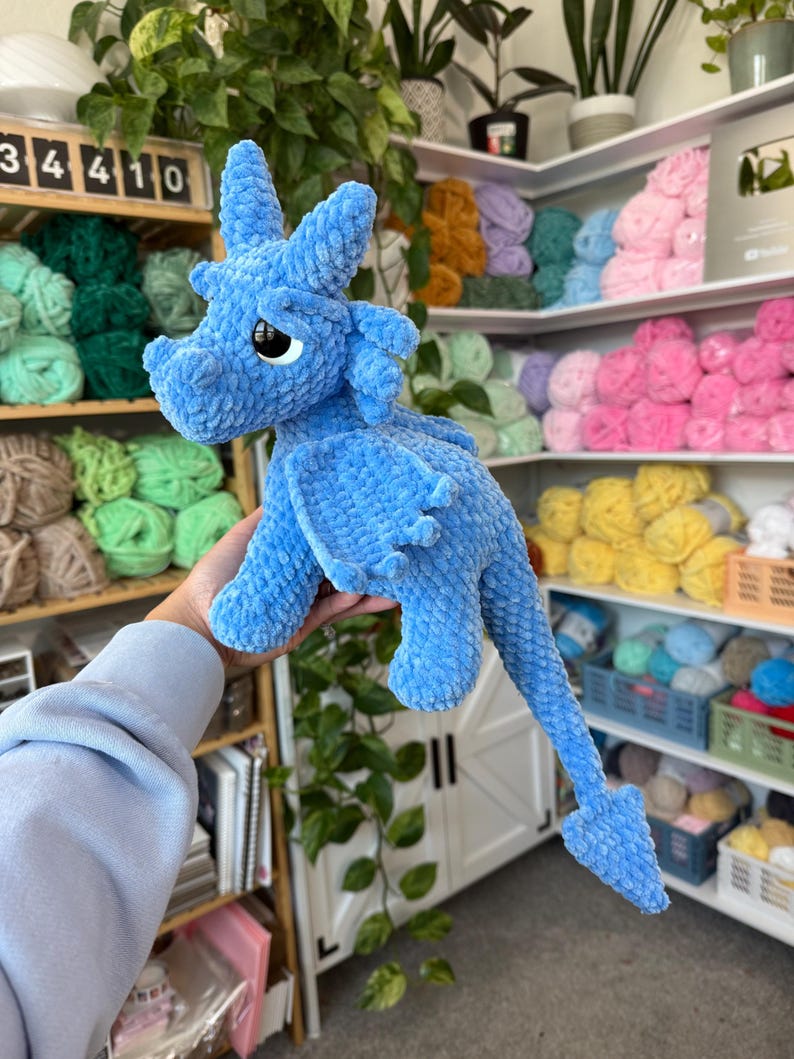K&ouml;nnte beinhalten: Ein hellblauer geh&auml;kelter Drache mit gro&szlig;en schwarzen Augen, Fl&uuml;geln und einem langen Schwanz. Der Drache ist aus einem weichen, strukturierten Garn gefertigt. Der Hintergrund zeigt Regale voller bunter Wolle.