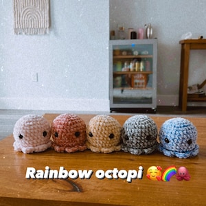 Op de afbeelding: Vijf gehaakte octopus knuffels in een regenboog van kleuren, roze, rood, geel, grijs en blauw, zijn op een houten tafel uitgelijnd. De tekst "Rainbow octopi" staat op de tafel voor de knuffels.