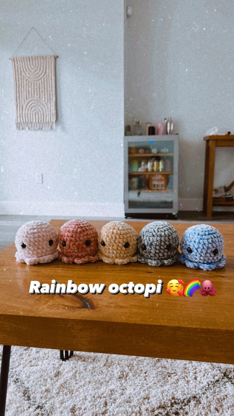 Op de afbeelding: Vijf gehaakte octopus knuffels in een regenboog van kleuren, roze, rood, geel, grijs en blauw, zijn op een houten tafel uitgelijnd. De tekst "Rainbow octopi" staat op de tafel voor de knuffels.