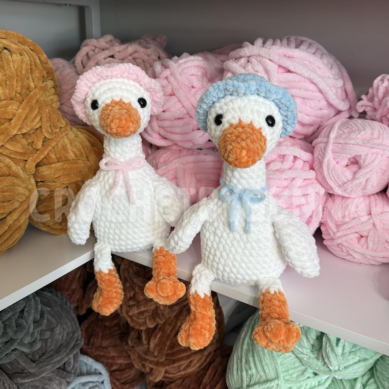 May include: Two handmade crochet plush geese with white bodies and orange beaks and feet. One goose wears a light pink bonnet and matching neck ribbon, while the other wears a light blue bonnet and matching neck ribbon. Both have black eyes.