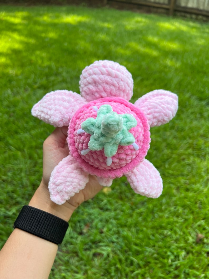 May include: A pink crocheted turtle with a green crocheted strawberry on its back.
