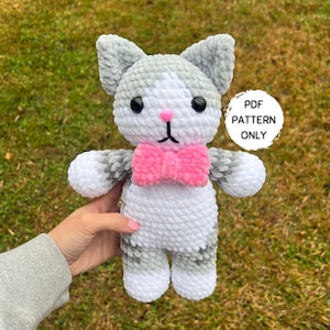Könnte beinhalten: Eine gehäkelte graue und weiße Katze mit einer rosa Schleife. Die Katze hält ein Schild mit der Aufschrift "PDF PATTERN ONLY".