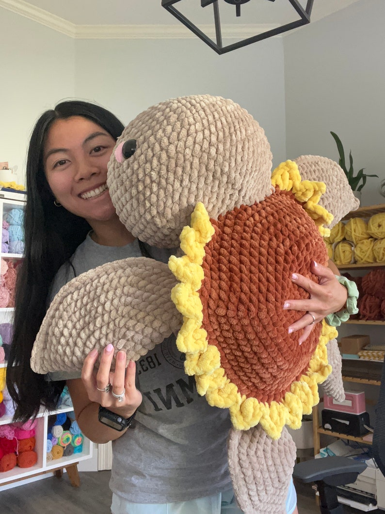 May include: A woman is holding a large crocheted turtle plush toy. The turtle is brown and tan with yellow accents. The woman is smiling and holding the turtle close to her.