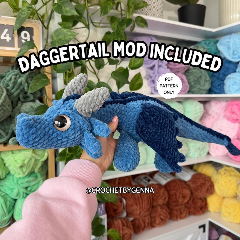 May include: A blue crocheted dragon with a daggertail mod included. The dragon is being held by a hand. The image also includes a sign that says "PDF Pattern Only".