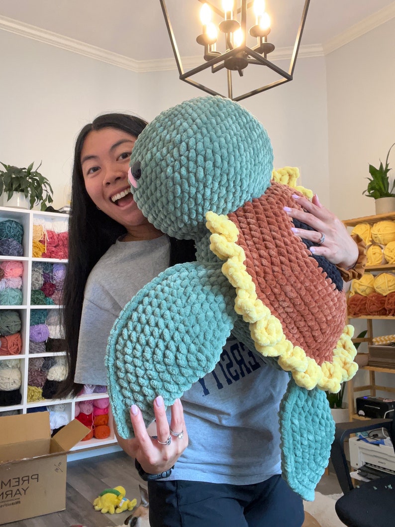 May include: A woman is holding a large crocheted sea turtle plush toy. The turtle is green, brown, and yellow. The woman is smiling and looking at the camera.