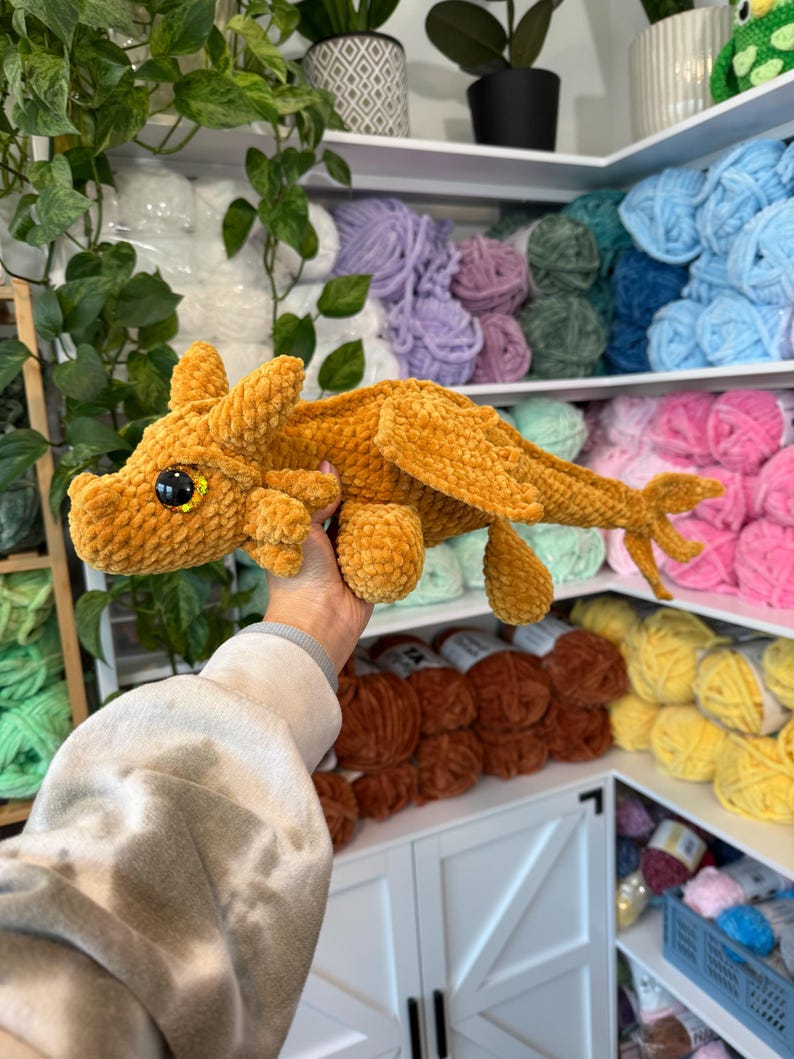 May include: A crocheted plush dragon toy in a golden brown color with large black eyes and a yellow iris. The dragon is being held by a hand in front of a shelf with colorful yarn.