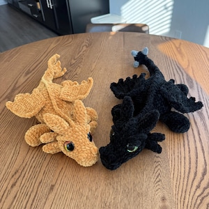 May include: Two crocheted dragon plushies, one brown and one black, with green eyes. The dragons are lying on a wooden table.
