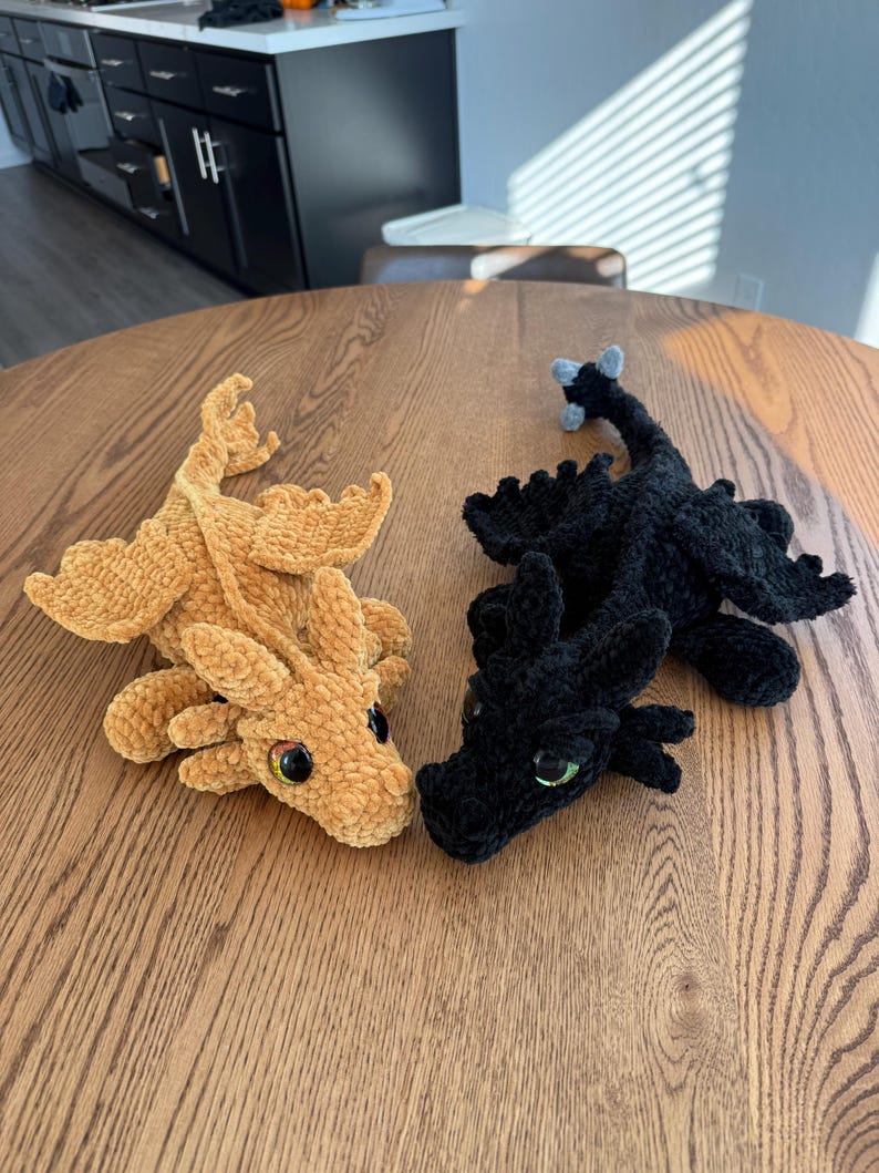 May include: Two crocheted dragon plushies, one brown and one black, with green eyes. The dragons are lying on a wooden table.