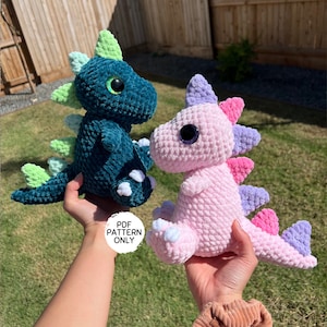 Puede incluir: Dos peluches de dinosaurio de crochet, uno verde azulado y otro rosa. El dinosaurio verde azulado tiene p&uacute;as verdes y garras blancas. El dinosaurio rosa tiene p&uacute;as moradas, rosas y blancas. Ambos dinosaurios tienen ojos grandes y negros.