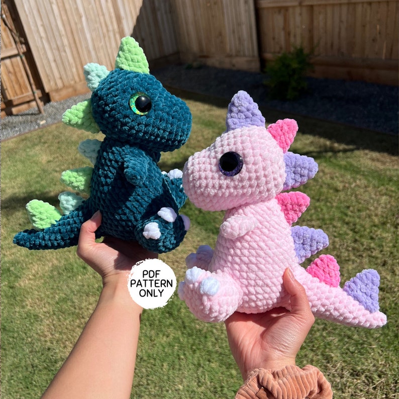 Puede incluir: Dos peluches de dinosaurio de crochet, uno verde azulado y otro rosa. El dinosaurio verde azulado tiene p&uacute;as verdes y garras blancas. El dinosaurio rosa tiene p&uacute;as moradas, rosas y blancas. Ambos dinosaurios tienen ojos grandes y negros.