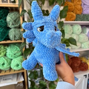 K&ouml;nnte beinhalten: Ein hellblauer geh&auml;kelter Drache mit gro&szlig;en, dunklen Augen und kleinen Fl&uuml;geln. Das Pl&uuml;schtier hat ein strukturiertes, flauschiges Aussehen. Der Drache wird vor einem Regal mit verschiedenen Garnfarben gehalten.
