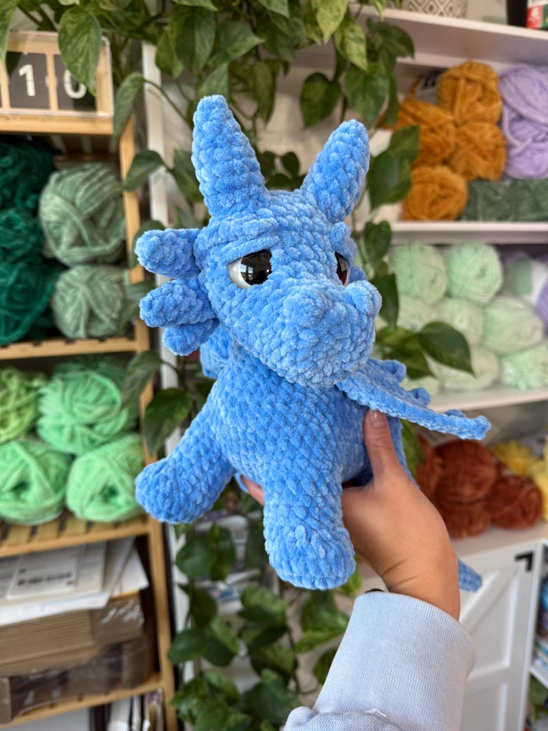 K&ouml;nnte beinhalten: Ein hellblauer geh&auml;kelter Drache mit gro&szlig;en, dunklen Augen und kleinen Fl&uuml;geln. Das Pl&uuml;schtier hat ein strukturiertes, flauschiges Aussehen. Der Drache wird vor einem Regal mit verschiedenen Garnfarben gehalten.