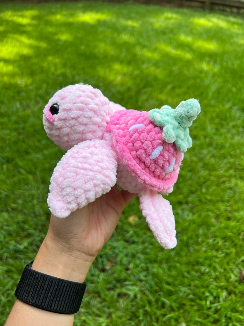 May include: A pink crocheted turtle with a strawberry shell. The turtle has a green leaf on top of the shell.