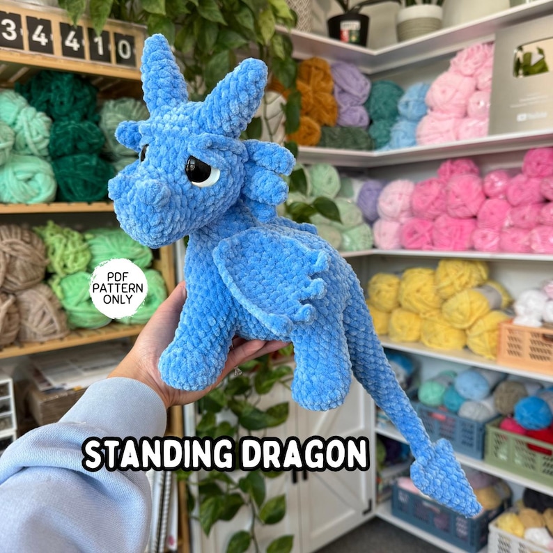K&ouml;nnte beinhalten: Ein hellblauer, stehender Pl&uuml;schdrache mit gro&szlig;en schwarzen Augen, Fl&uuml;geln und einem langen Schwanz. Der Drache besteht aus weichem, strukturiertem Material. Der Text "STANDING DRAGON" wird am unteren Bildrand angezeigt.