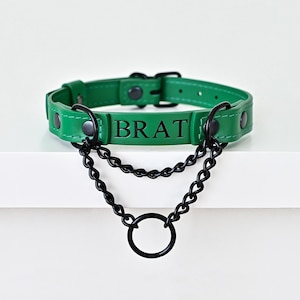 May include: Green leather collar with the word "BRAT" in black lettering. The collar has black metal hardware and a double chain with a ring. The collar is on a white surface.