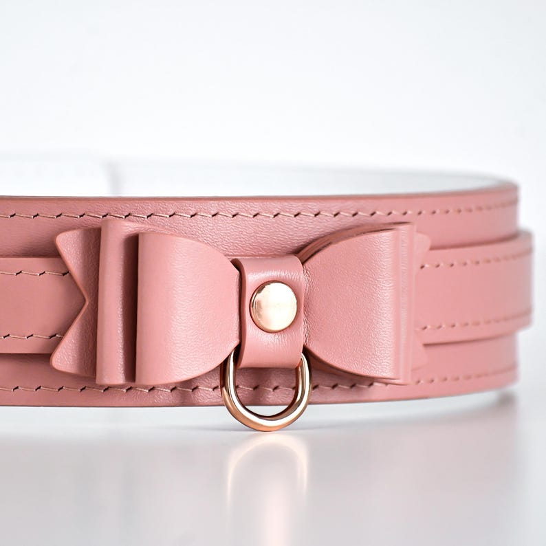 Pu&ograve; includere: Un collare in pelle rosa con un dettaglio a fiocco e un anello in metallo color oro. Il collare &egrave; cucito e ha un interno bianco. Il fiocco &egrave; centrato sul collare.