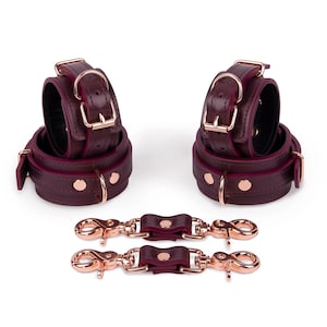 Può includere: Restrizioni per polsi in pelle bordeaux con finiture in oro rosa. Il set include due manette da polso e due cinghie di collegamento con moschettoni. I polsini hanno un interno imbottito e sono fissati con fibbie.