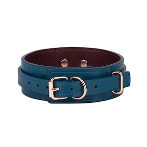 May include: A teal leather collar featuring rose gold hardware, including a buckle, a D-ring, and a keeper loop. The interior of the collar is lined with a dark burgundy leather, and the construction is secured with two visible rose gold rivets.