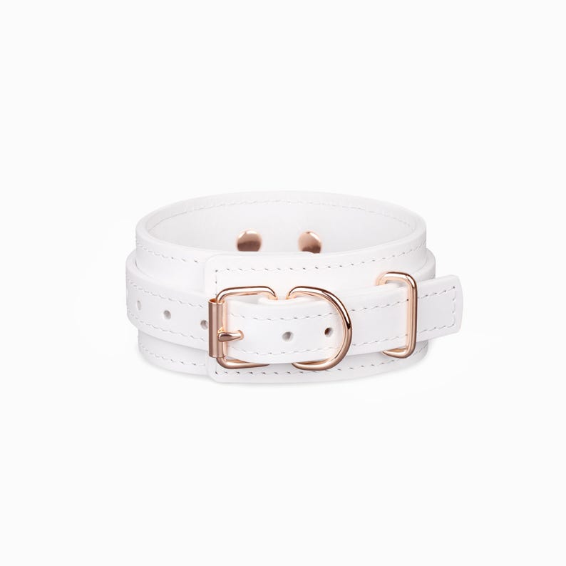 May include: A white leather-like cuff bracelet featuring a rose gold-toned metal buckle, a matching D-ring, and two small rose gold-toned rivets visible on the inner band. The accessory has a smooth finish with visible stitching along the edges.
