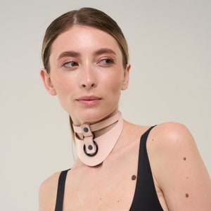 Może przedstawiać: Jasnoróżowy skórzany choker z czarnym metalowym pierścieniem i okuciami. Choker ma konstrukcję z podwójnym paskiem i zapięcie na guzik. Produkt jest noszony na szyi.