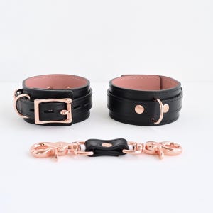 Op de afbeelding: Twee zwarte polsbanden met een zachte roze voering, voorzien van gespen, D-ringen en klinknagels in roségoudkleurig metaal. Een bijpassende zwarte verbindingsriem met draaibare karabijnhaken in roségoudkleurig metaal ligt ervoor.