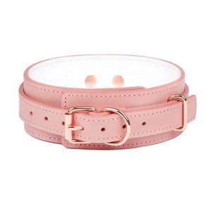 May include: A light pink collar featuring a white interior lining, rose gold-toned metal hardware, a buckle closure, and a D-ring. The collar has visible stitching along the edges and multiple adjustment holes for a secure fit around the neck.