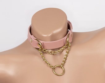 May include: A light pink faux leather choker collar featuring gold-toned metal hardware, including two layered chains and a central circular ring. The collar is secured with gold-toned rivets and an adjustable buckle closure on the side.