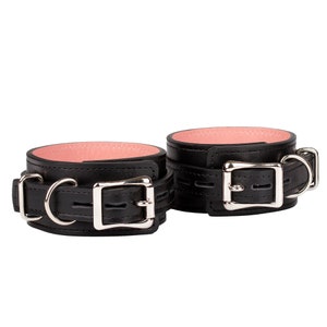 May include: A pair of black leather wrist restraints with a pink interior. Each restraint features a silver-colored buckle and a D-ring for secure fastening. The cuffs are designed with a classic, minimalist aesthetic.