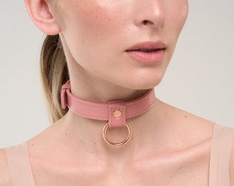 May include: A pink leather choker necklace with a gold-colored ring and buckle closure. The choker is a solid color and has a simple, elegant design. The ring is attached to the center of the choker.
