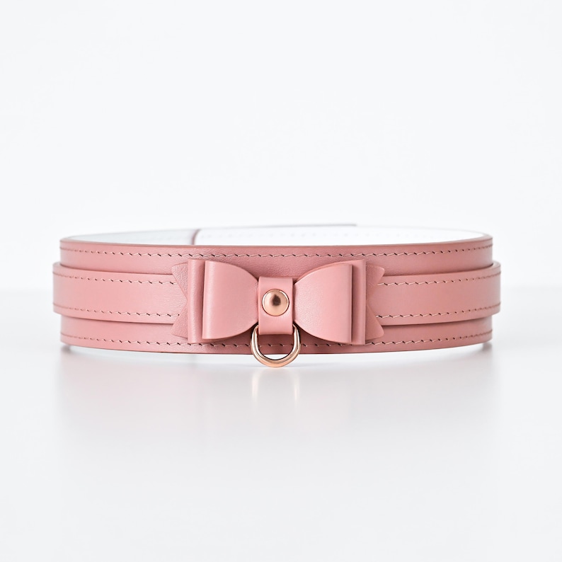 Pu&ograve; includere: Un collare in pelle rosa con un dettaglio a fiocco e un anello in metallo color oro. Il collare ha un design a strati con cuciture visibili e un interno bianco. Il fiocco &egrave; centrato sul davanti.