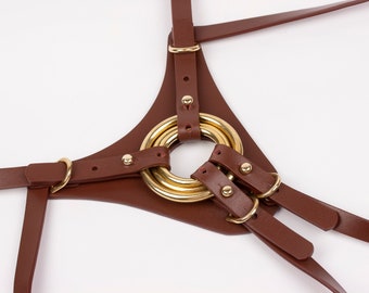 Puede incluir: Un arnés de cuero marrón con un panel central triangular conectado a varias correas ajustables. El diseño incorpora anillos y remaches de metal en tono dorado pulido, con las correas aseguradas mediante pequeñas hebillas metálicas.
