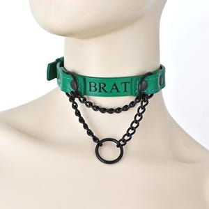 May include: A green choker collar featuring the word BRAT printed in black letters. The accessory includes two black metal chains hanging from the band, connected to a central black metal ring. The band is secured with black metal rivets.