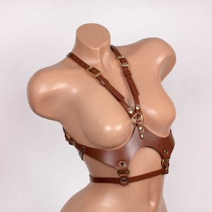 Op de afbeelding: Bruin leren lichaamsharnas met verstelbare bandjes en goudkleurige hardware. Het harnas heeft een halter-stijl halslijn en een borstontwerp met een uitsparing. De bandjes zijn verbonden met metalen ringen en gespen.