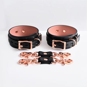 Könnte beinhalten: Zwei schwarze Handgelenksmanschetten mit weichem, rosafarbenem Innenfutter, ausgestattet mit Schnallen, D-Ringen und Nieten in Roségold-Optik. Enthalten sind zwei passende schwarze Verbindungsriemen mit drehbaren Karabinerhaken in Roségold.
