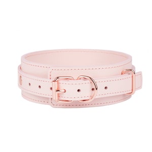 May include: A pale pink collar featuring a wide band design with rose gold-toned metal hardware, including a buckle, a D-ring, and a rectangular loop. The collar has visible stitching along the edges and multiple adjustment holes for sizing.