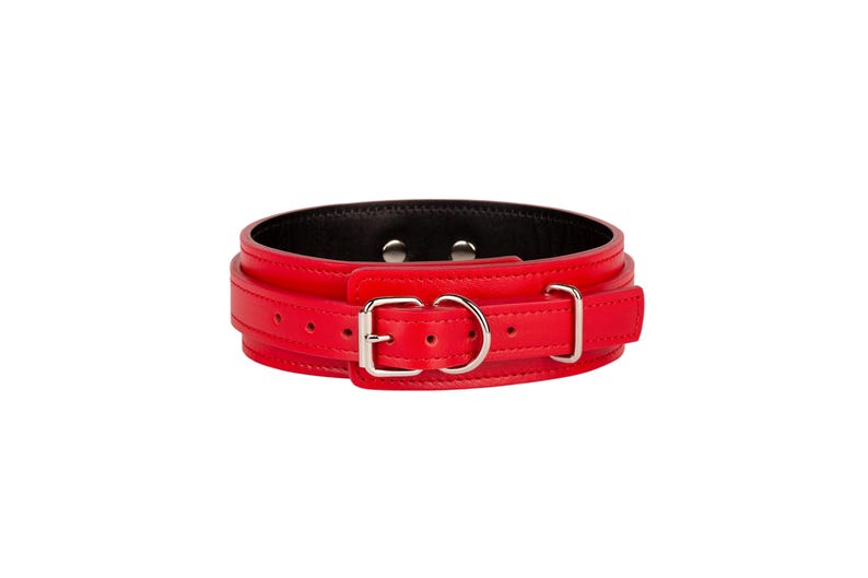 May include: A bright red collar featuring a smooth exterior finish, a black interior lining, and silver-toned metal hardware including a buckle, a D-ring, and two rivets. The collar is constructed with a wide band and a narrower adjustable strap.