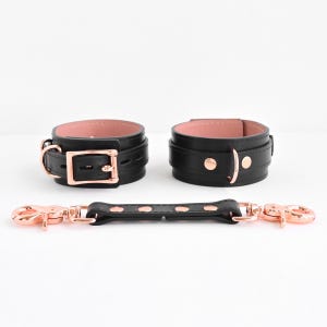 Könnte beinhalten: Ein Set aus zwei schwarzen Leder-Manschetten mit weichem, rosafarbenem Innenfutter und roségoldenen Beschlägen, inklusive eines passenden Verbindungsriemens aus schwarzem Leder mit roségoldenen Karabinerhaken an beiden Enden.