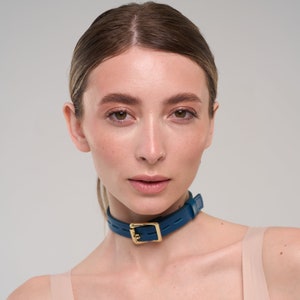 Puede incluir: Un collar de gargantilla de cuero color turquesa con una hebilla dorada. La gargantilla es una banda simple con una hebilla rectangular. La modelo lleva la gargantilla alrededor del cuello.