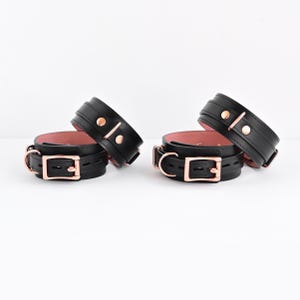 May include: A pair of black wrist cuffs with a soft pink interior lining. Each cuff features rose gold-toned metal hardware, including a rectangular buckle, a D-ring, and two circular studs. The cuffs are adjustable with multiple pre-cut holes.