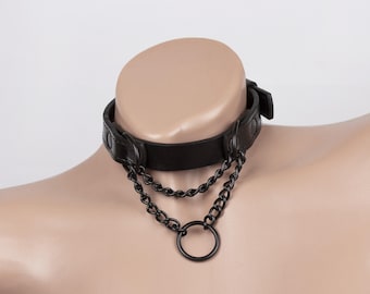 Può includere: Un collare nero caratterizzato da una fascia liscia con dettagli a borchie circolari. Due catene in metallo nero pendono dalla parte anteriore, collegandosi a un anello circolare centrale. Il collare include una chiusura con fibbia laterale.