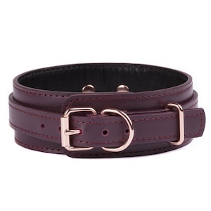 May include: A deep burgundy leather collar featuring rose gold-toned metal hardware, including a rectangular buckle, a D-ring, and a sliding keeper loop. The collar has visible tonal stitching along the edges and a smooth, dark interior lining.