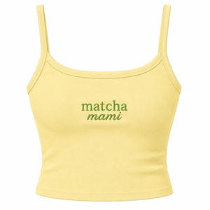 May include: A pale yellow sleeveless crop top with thin shoulder straps and a scoop neckline. The center of the chest features the text "matcha mami" embroidered in a dark green thread, with the word "matcha" in a serif font and "mami" in italics.