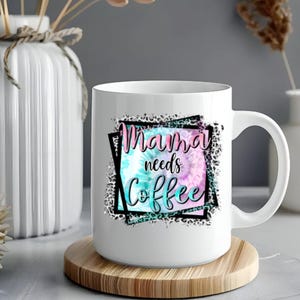 May include: White ceramic coffee mug with the text "Mama needs Coffee" in a tie-dye design of pink, blue, and teal. The mug sits on a wooden coaster. A white vase and decorative items are in the background.