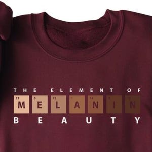 May include: A maroon sweatshirt with the phrase "THE ELEMENT OF MELANIN BEAUTY" printed on the front. The text is in white, with the word "MELANIN" represented by a series of skin tone color blocks.