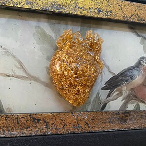 Op de afbeelding: Een gouden hartvormig harsbeeld met bladgoudaccenten, ingelijst in een zwart en gouden lijst.